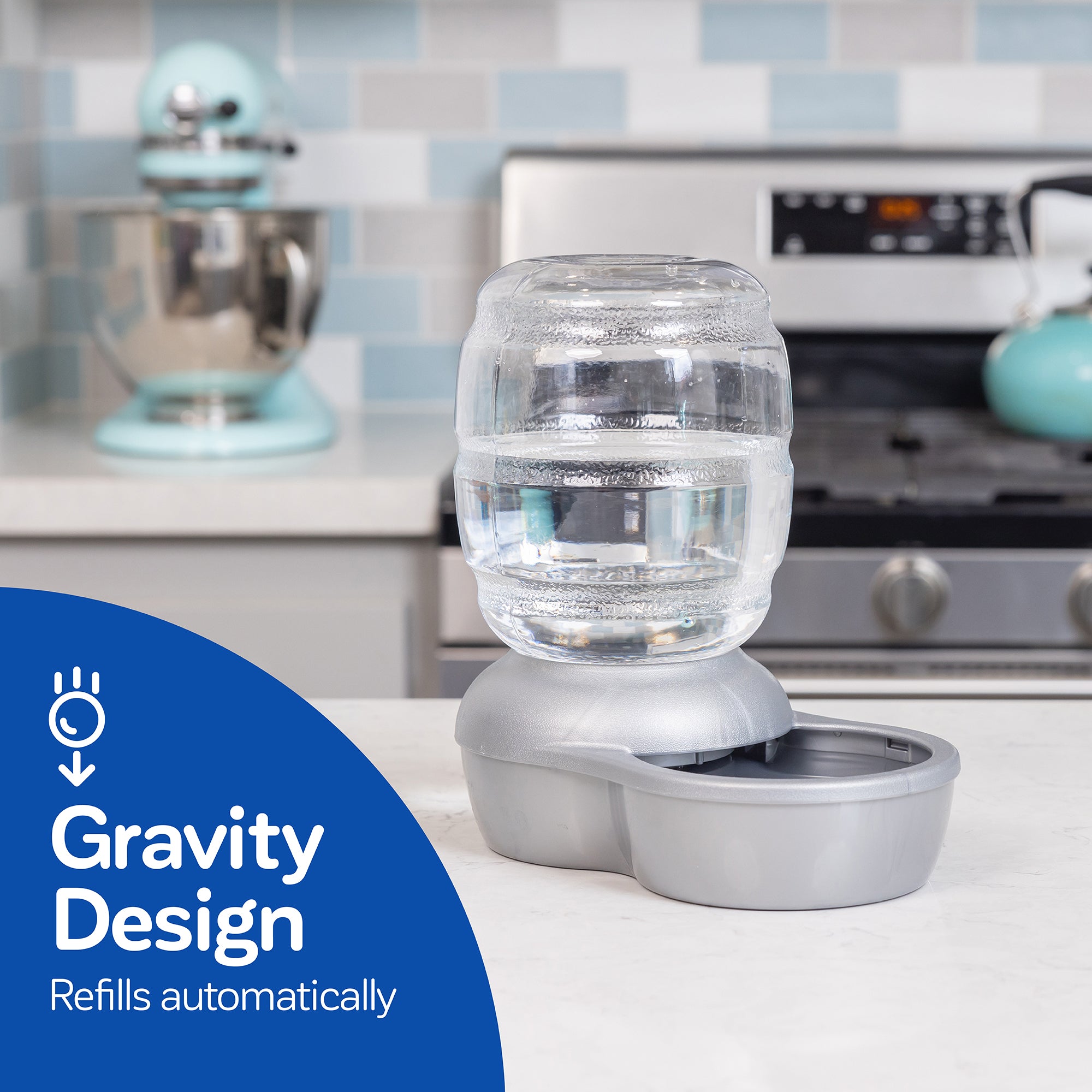 On the kitchen counter sits a Pet-mates Replendish Pet Waterer with Microban, featuring a clear jug and blue circle labeled Gravity Design Refills Automatically. It keeps pet water fresher while the tiled backsplash and appliances can be seen in the background.