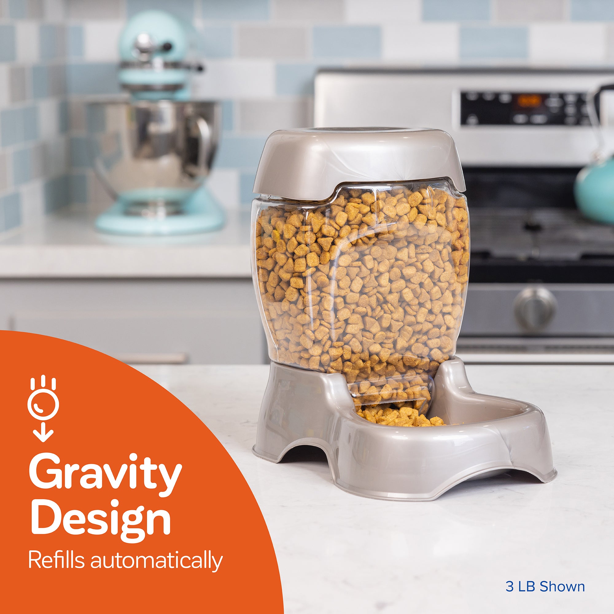 A Pet-mates Pet Cafe Feeder with a clear hopper filled with dry kibble sits on the kitchen counter, ensuring automatic refills. Behind it are a mixer and stove, accompanied by an orange graphic stating Gravity Design Refills Automatically.
