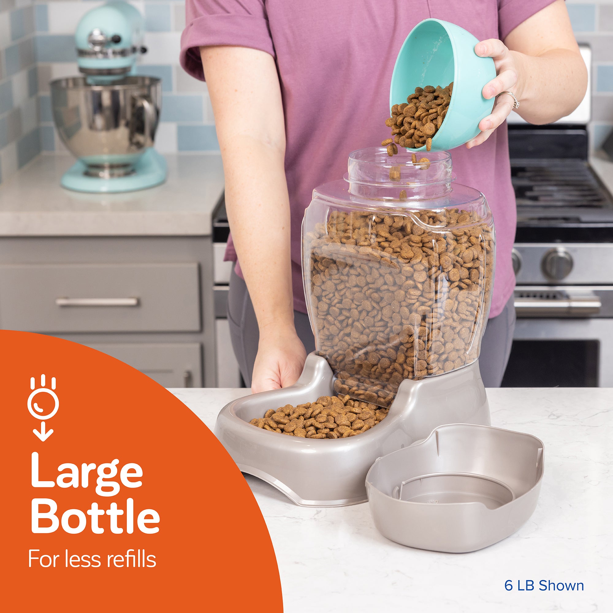 A person pours pet food from a teal bowl into the Pet-mates Pet Cafe Feeder, which is a large transparent gravity feeder with an attached gray dish for reduced refill frequency, sitting on the kitchen counter. In the background, theres a mixer and a stove. 6 LB Shown.