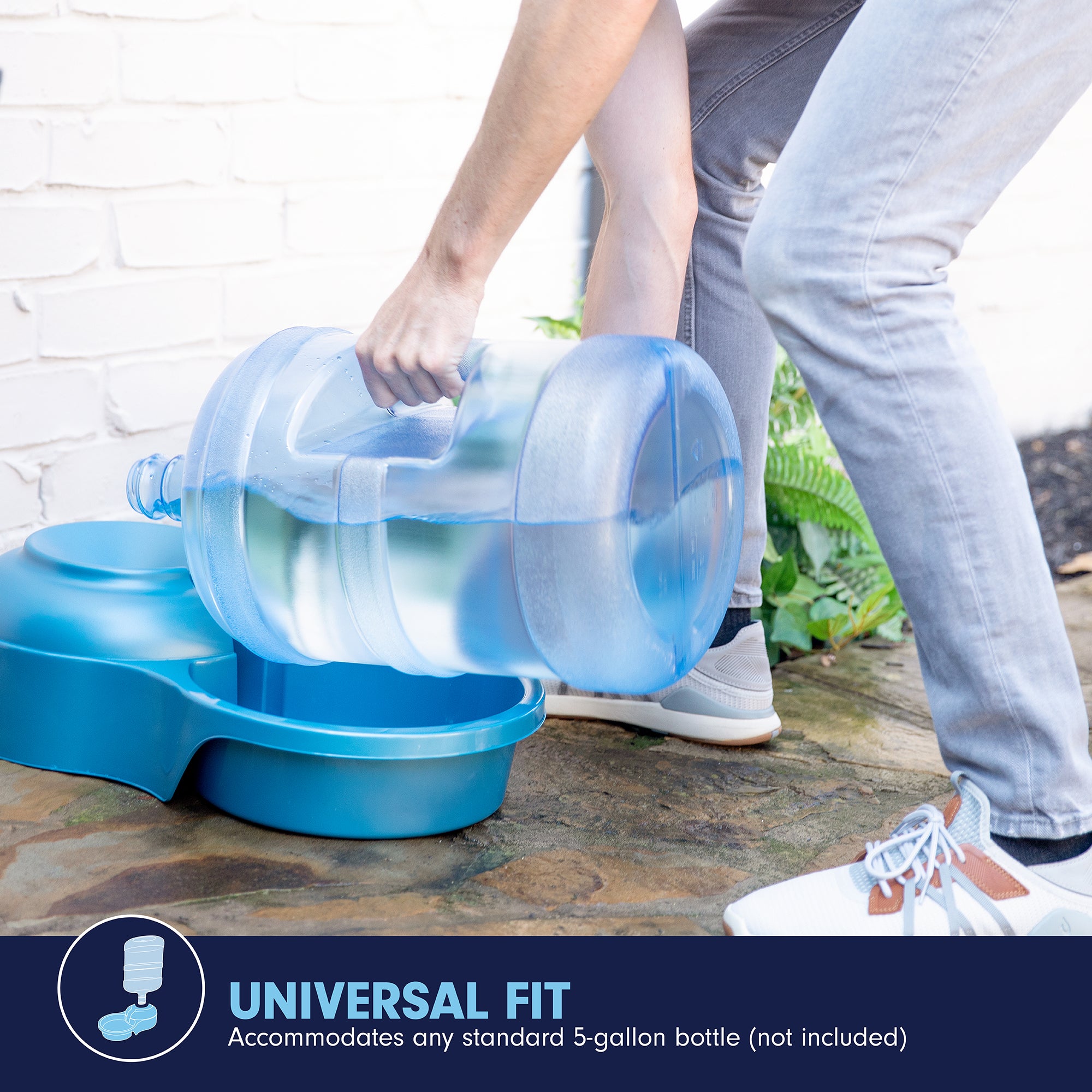 Wearing jeans and sneakers, a person places a large clear 5-gallon water bottle into the Pet-mates Big Blue Bowl, a blue pet water dispenser. Perfect for outdoor hydration, it accommodates any standard 5-gallon bottle (not included), ensuring pets always have fresh water.