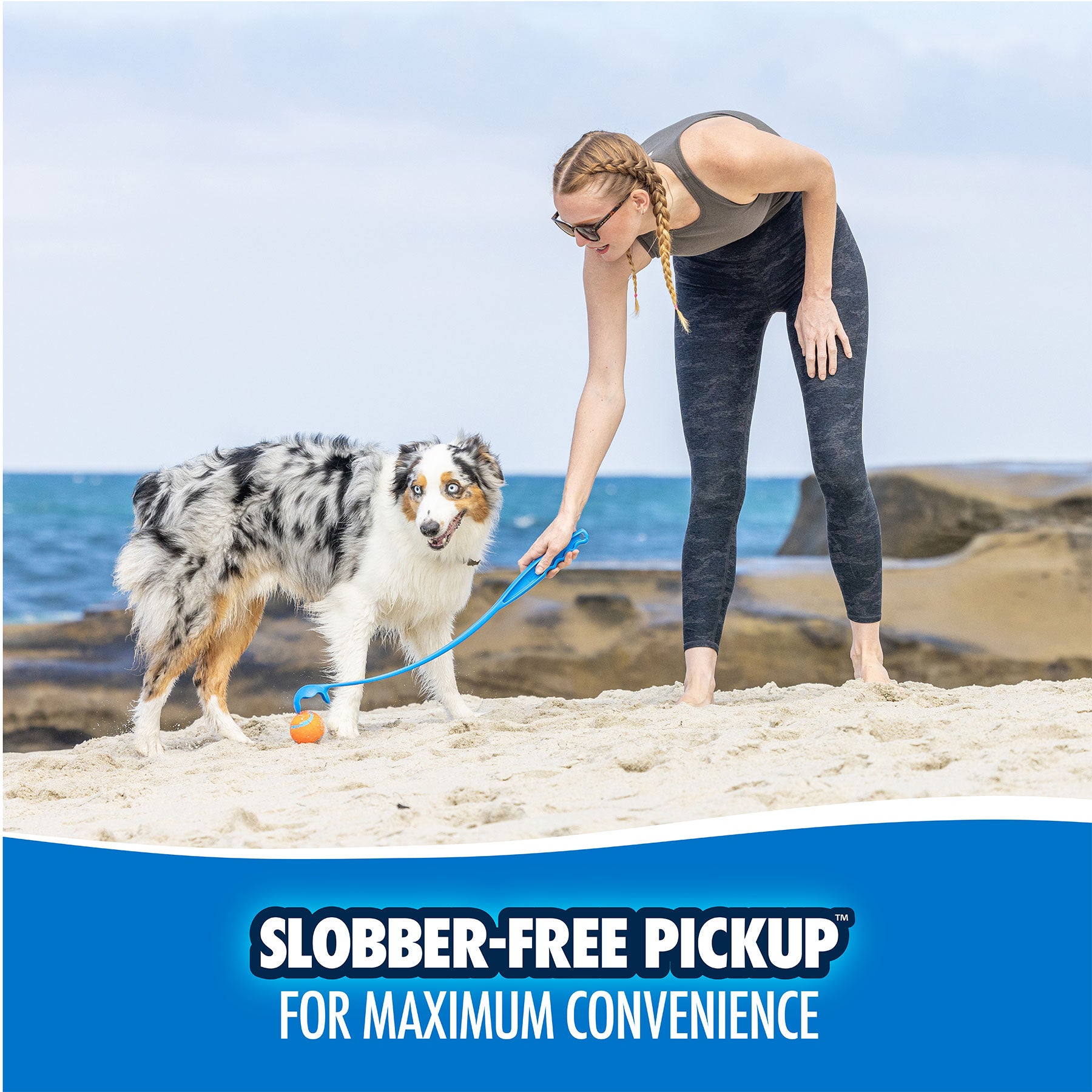 A woman with braided hair uses the Chuckit Amphibious Fetch Pack to grab an orange ball while her fluffy dog stands on a sandy beach. The ocean and rocks are visible behind them. Text reads, SLOBBER-FREE PICKUP FOR MAXIMUM CONVENIENCE.