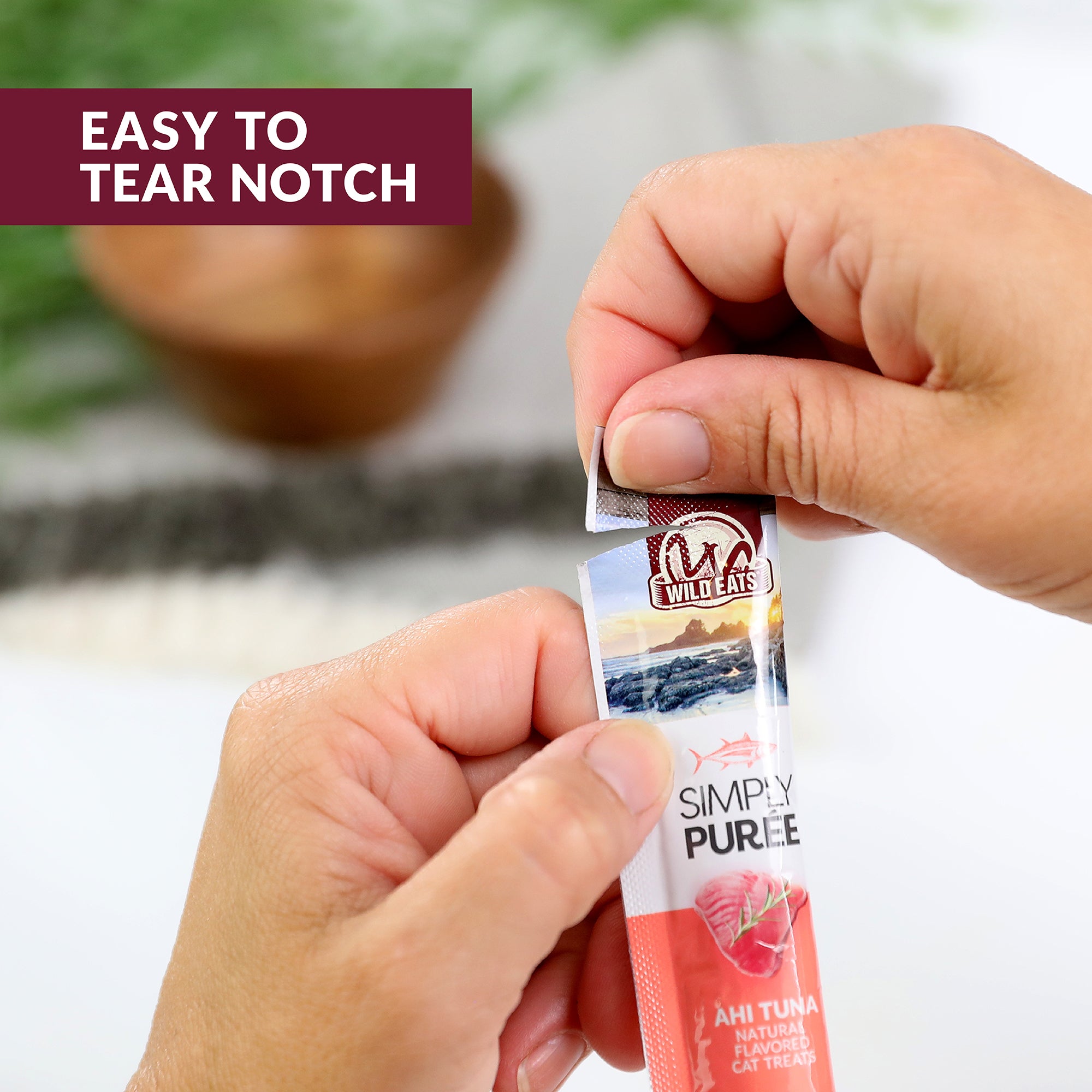 A hand opens a Wild Eats Simply Puree Variety Pack cat treat. The design features an Easy to Tear Notch in the top left corner. This natural pet food, rich in Omega-3 for cat hydration, is set against a blurred plant and bowl background.