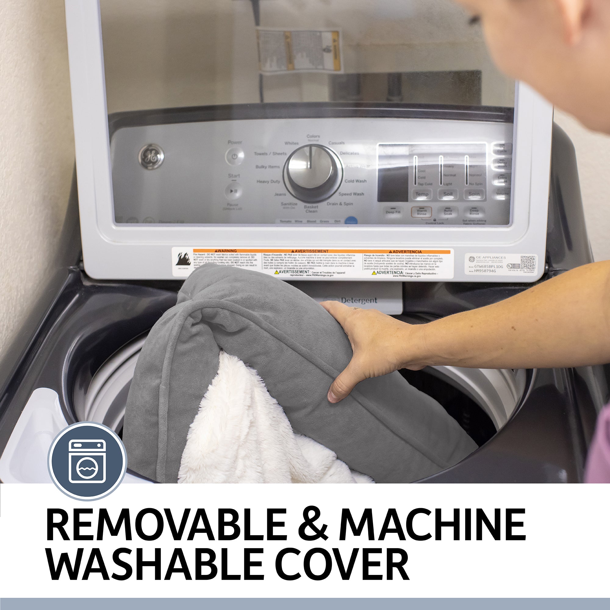 A person places the gray PetFusion Premium Reversible Micro Plush Dog Blanket into the washing machine. The washer’s panel shows REMOVABLE & MACHINE WASHABLE COVER, emphasizing its polyester micro plush design as a furniture protector.