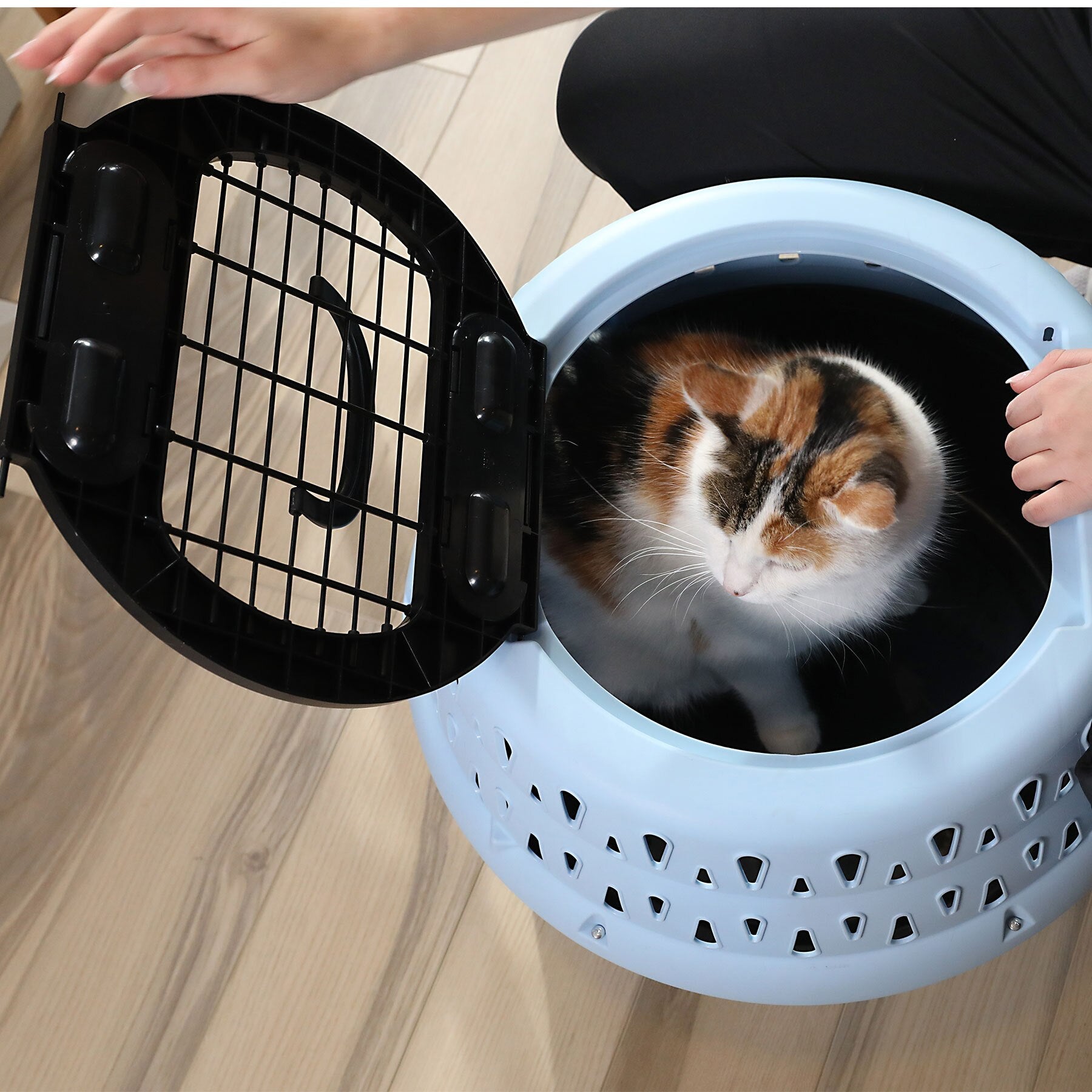 A calico cat relaxes inside the Pet-mates Get-A-Round E-Z Load Kennel For Cats, enjoying a secure, comfortable spot as a person kneels nearby on the light wooden floor.