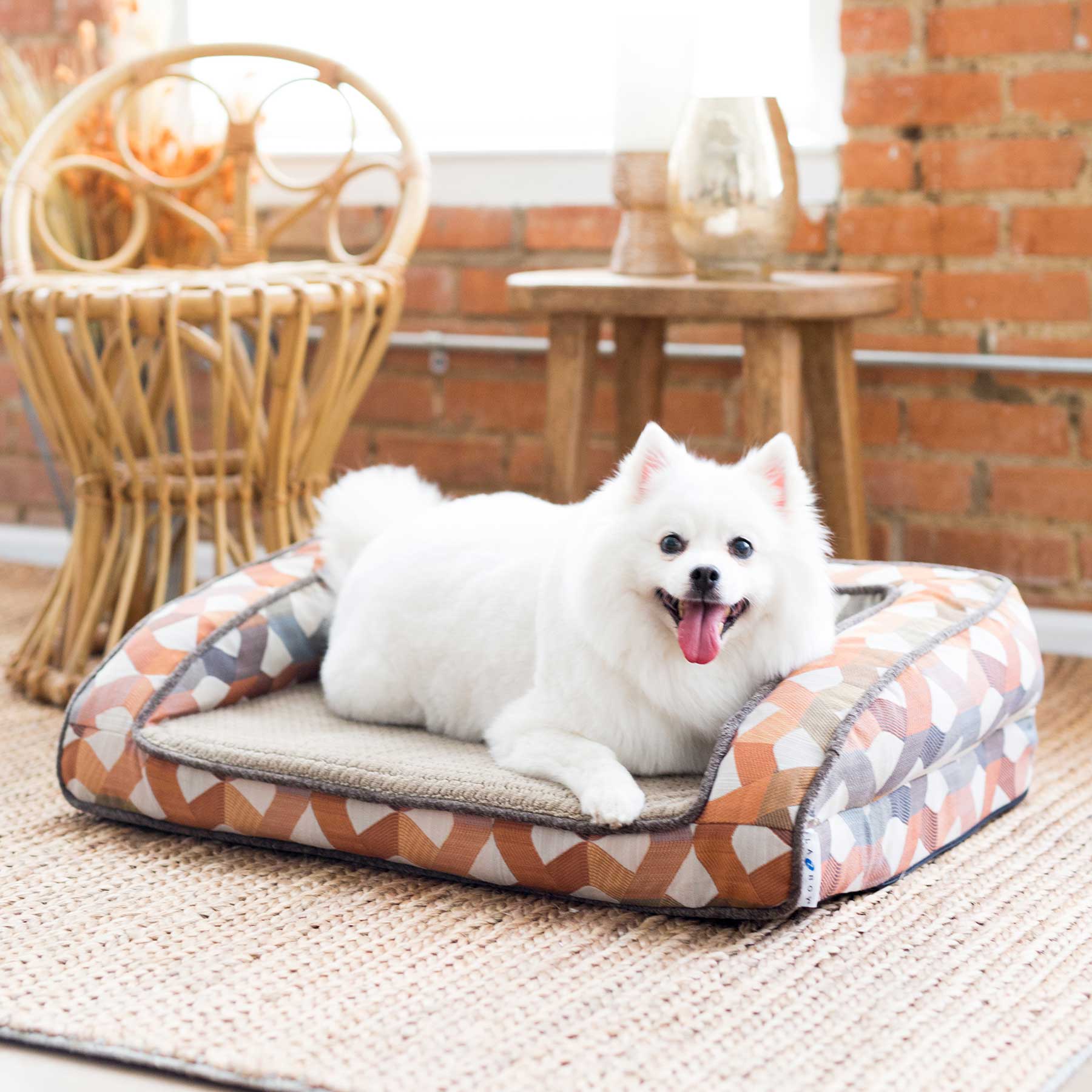 A fluffy white dog basks in comfort on a La-Z-Boy Riley Orthopedic Sofa Dog Bed featuring Gel Memory Foam and a beige cushion. Its tongue lolls happily as it relaxes. A wicker chair and wooden stool sit against a brick wall, enhancing the cozy atmosphere.