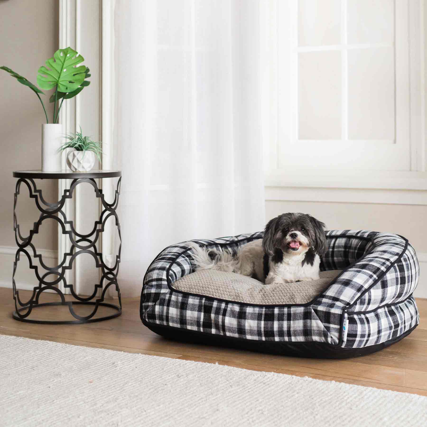 A small black and white dog relaxes in a La-Z-Boy Spencer Plaid Tucker Sofa Dog Bed, designed for medium-sized dogs, on a wooden floor next to a round metal side table with a potted plant. Natural light filters through tall windows with sheer curtains, creating a serene atmosphere.