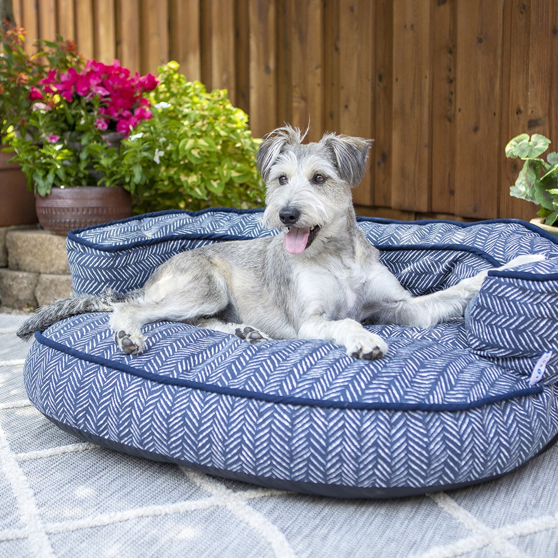 A happy dog with shaggy fur lounges on a La-Z-Boy Gatson Indoor Outdoor Dog Bed in blue pattern. Lush green plants, pink flowers, and a wooden fence create a calm and inviting outdoor setting.